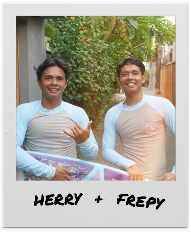 Herry and Frepy in their rashguards with a surfboard, smiling, one throwing a shaka